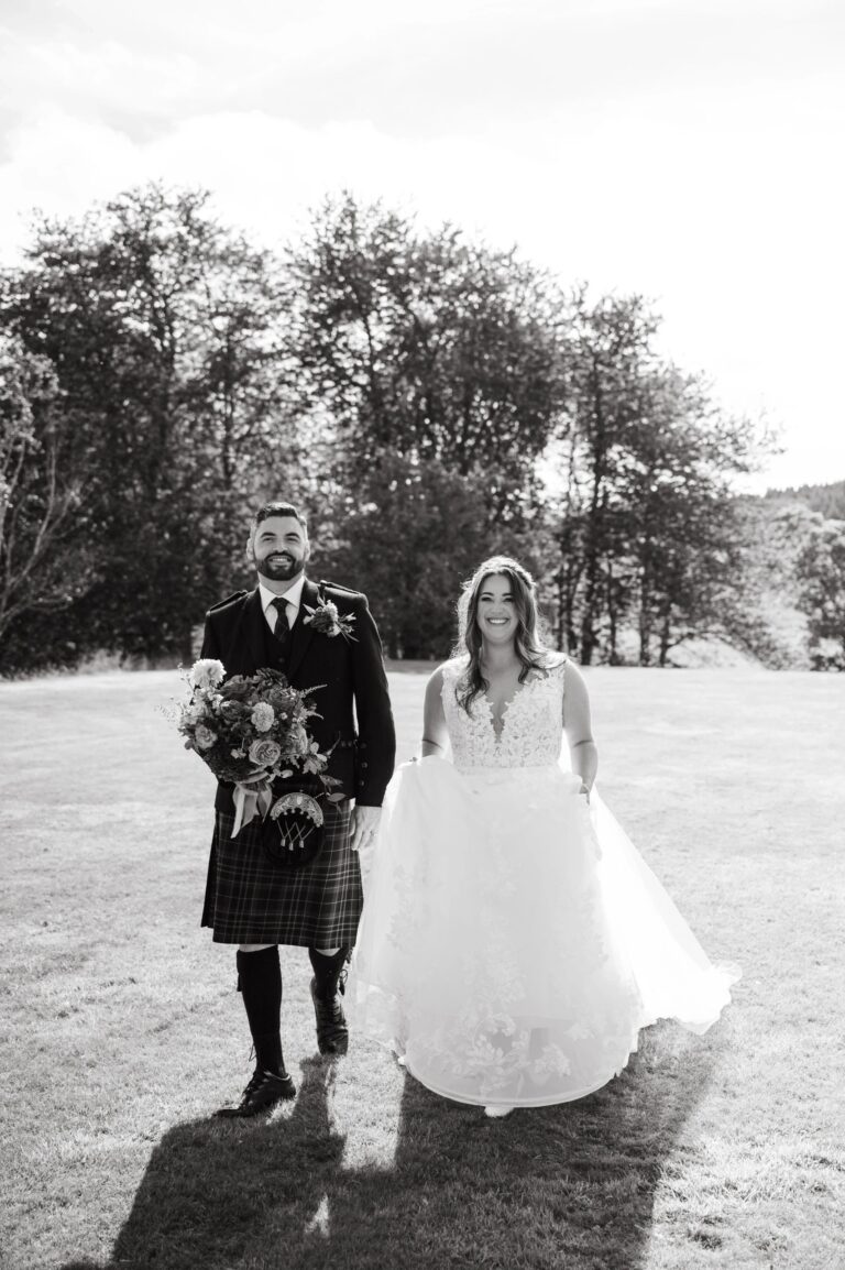 Bride and groom walking across the sunny grounds of Ballogie House wedding venue, groom in full Highland dress carrying the bridal bouquet, with rolling Scottish countryside in the background.