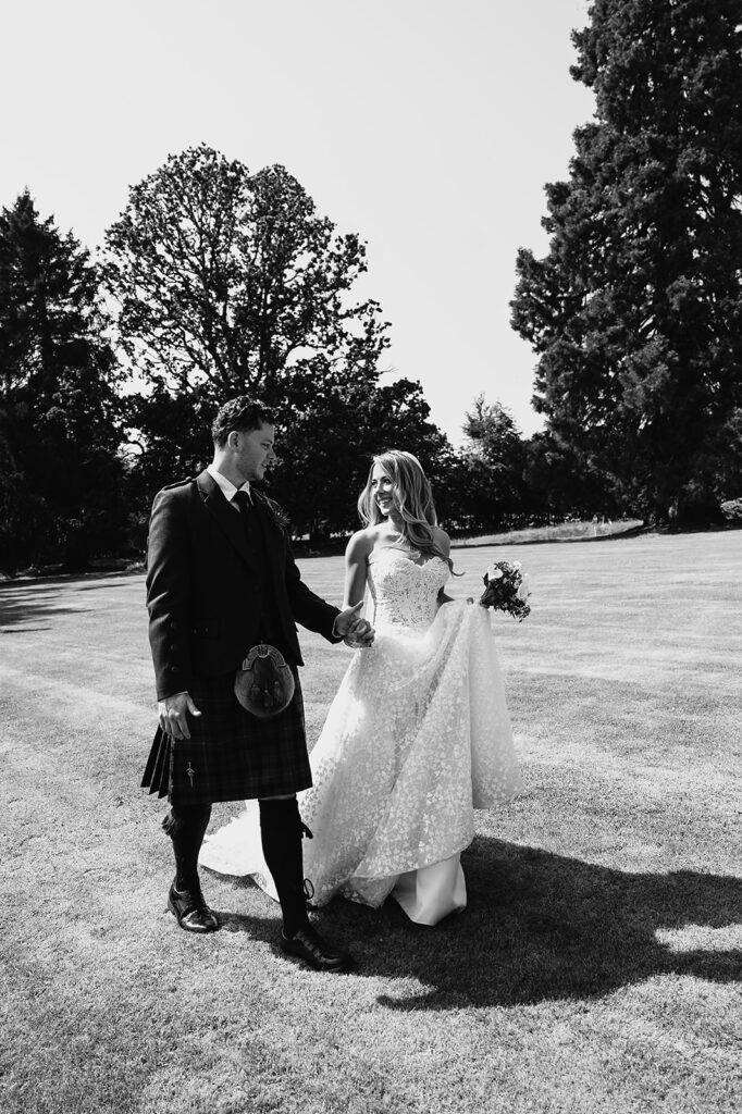 Sarah and Harry walking hand in hand at their Raemoir House wedding, bride in lace dress, groom in traditional tartan kilt